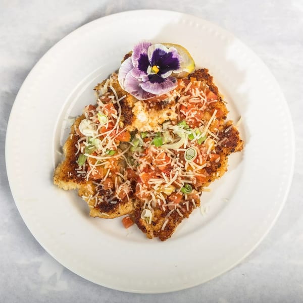 Crispy fried chicken cutlet topped with diced tomatoes, shredded cheese, green onions, and an edible flower.