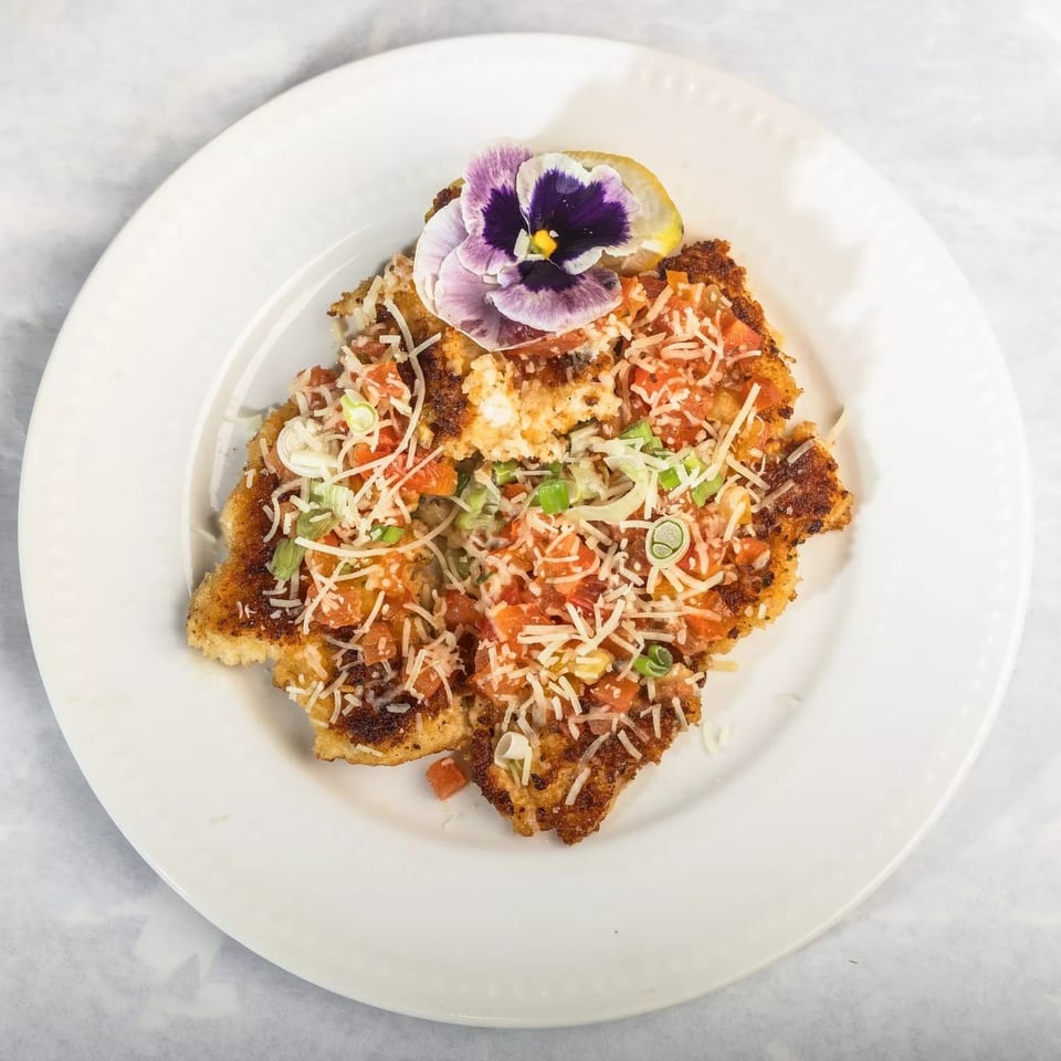 Crispy fried chicken cutlet topped with diced tomatoes, shredded cheese, green onions, and an edible flower.