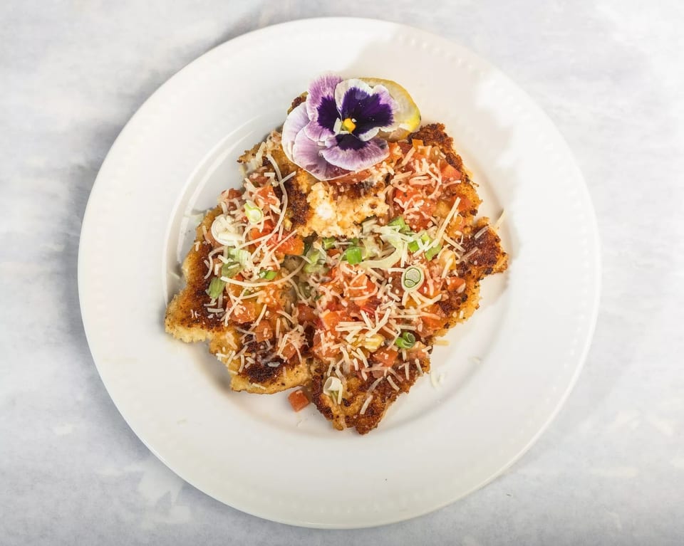 Crispy fried chicken cutlet topped with diced tomatoes, shredded cheese, green onions, and an edible flower.