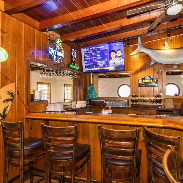 A rustic wooden bar with stools, neon signs, a TV, and a large fish decoration.