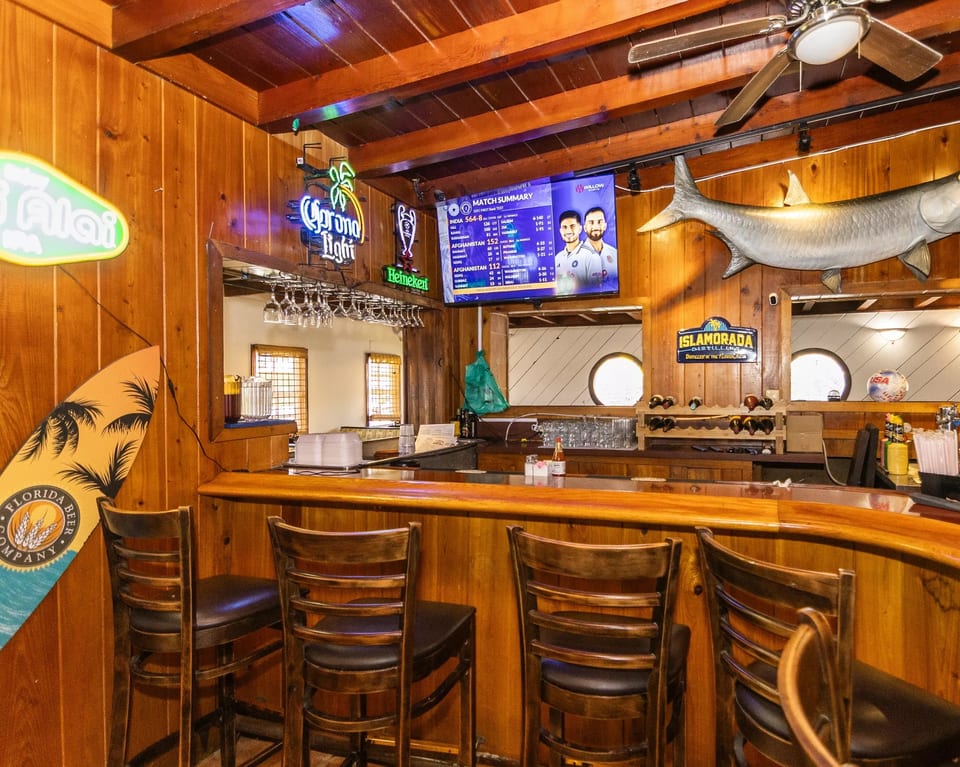 A rustic wooden bar with stools, neon signs, a TV, and a large fish decoration.