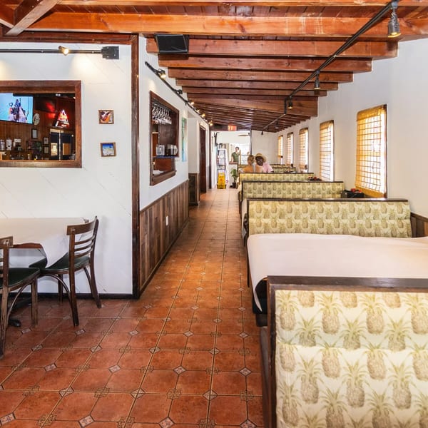 Restaurant interior with wooden beams, pineapple-patterned booths, tiled floor, and a bar pass-through.