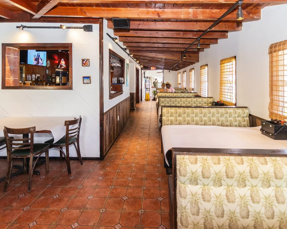 Restaurant interior with wooden beams, pineapple-patterned booths, tiled floor, and a bar pass-through.