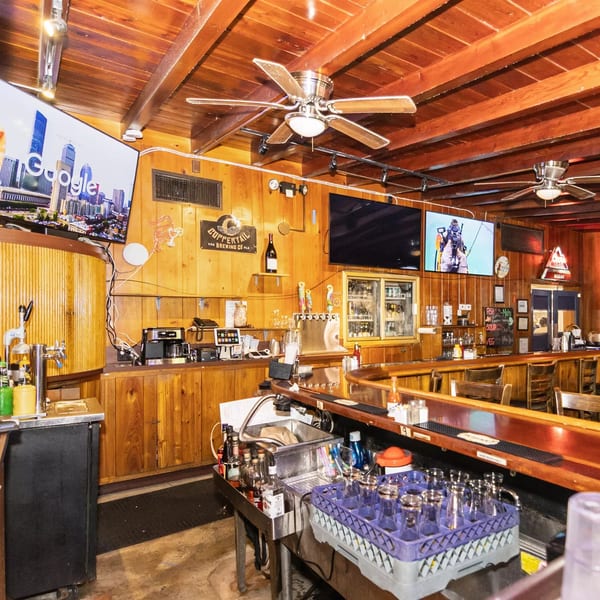 A rustic bar interior with wooden walls and ceiling, multiple TVs, beer taps, and bar stools.