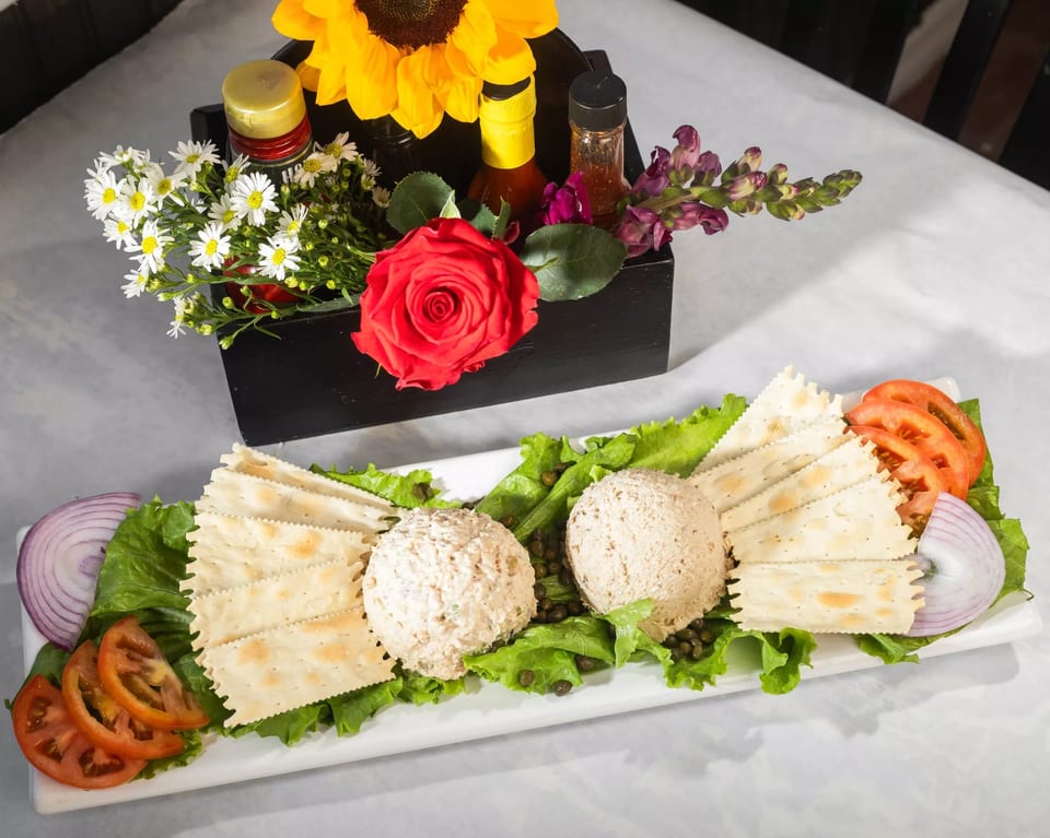 Two scoops of tuna salad with crackers, lettuce, tomatoes, and red onion, next to a floral centerpiece.