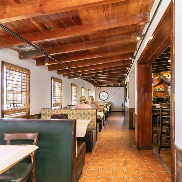 Long, narrow restaurant dining room with wooden beamed ceiling, booth seating, and tiled floor.