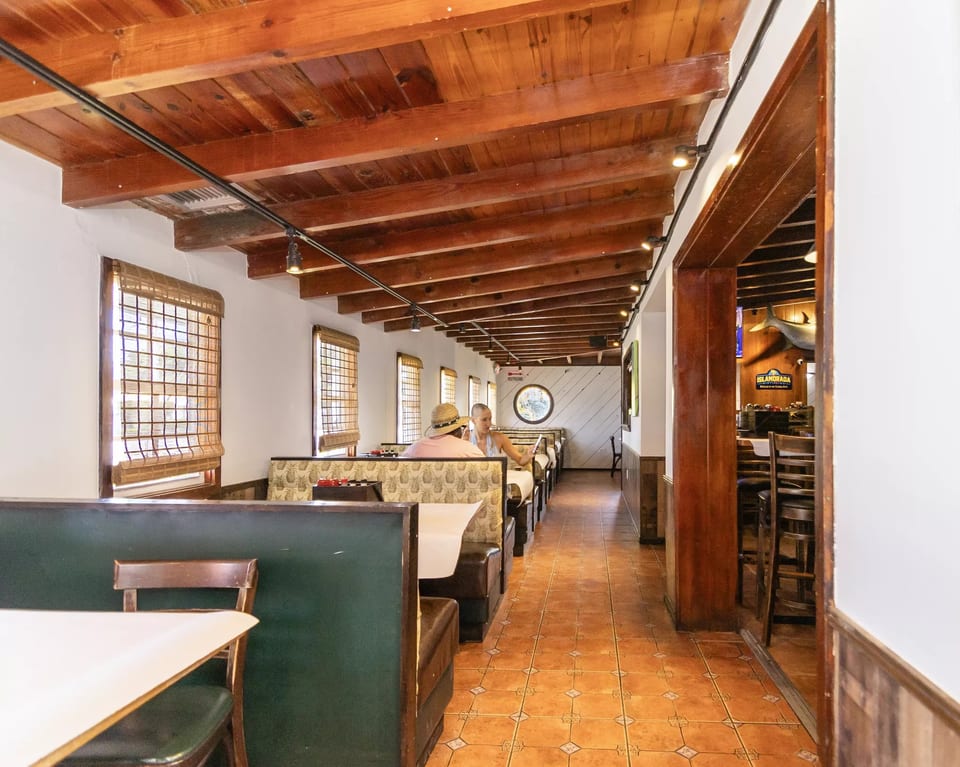 Long, narrow restaurant dining room with wooden beamed ceiling, booth seating, and tiled floor.