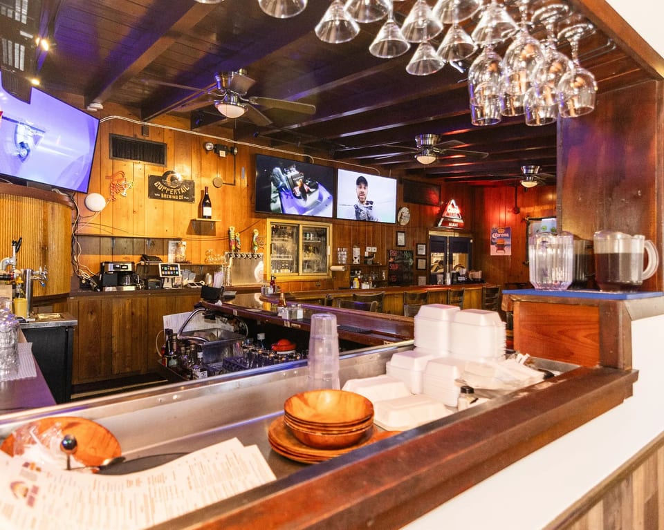 A rustic bar interior with wooden paneling, multiple TVs, beer taps, and hanging glassware.