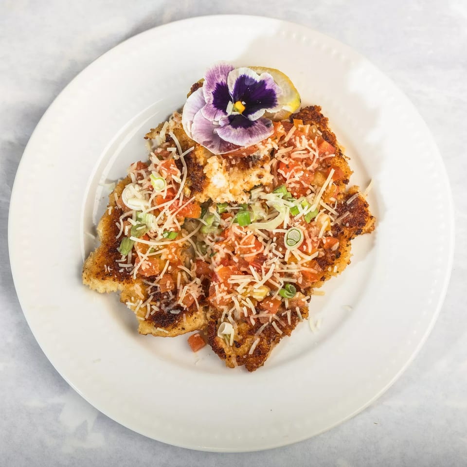 Crispy chicken Milanese topped with diced tomatoes, shredded cheese, green onions, and a purple edible flower.