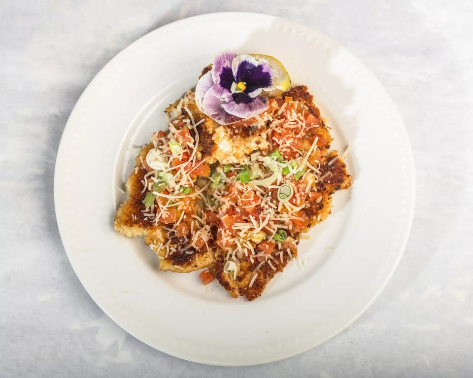 Crispy chicken Milanese topped with diced tomatoes, shredded cheese, green onions, and a purple edible flower.