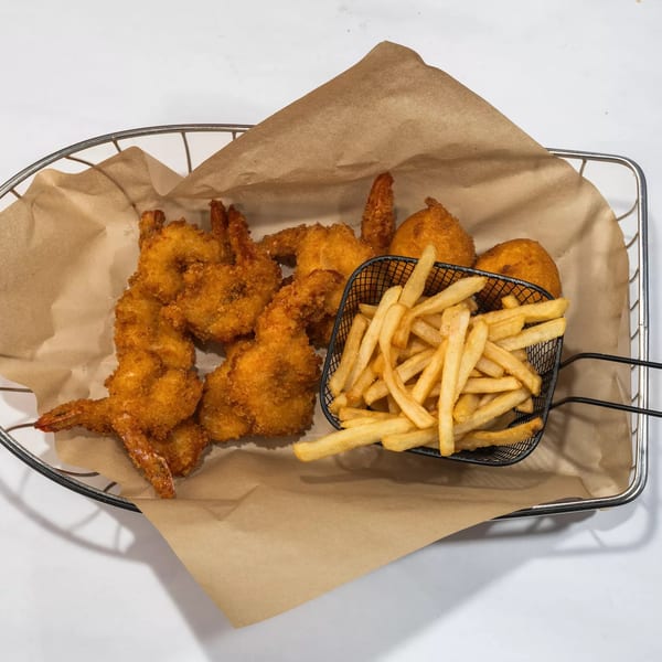 A metal basket with brown paper holds fried shrimp, hush puppies, and a mini basket of french fries.