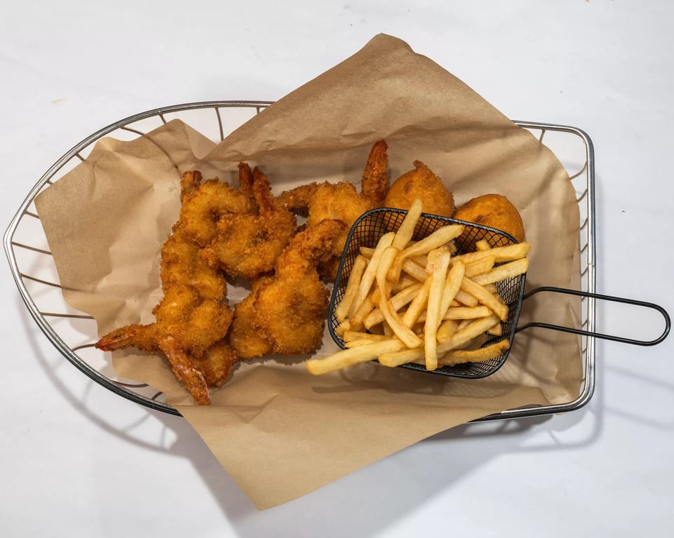 A metal basket with brown paper holds fried shrimp, hush puppies, and a mini basket of french fries.