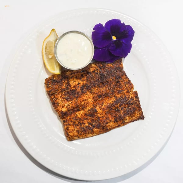 Blackened fish fillet with a lemon wedge, tartar sauce, and a purple edible flower on a white plate.