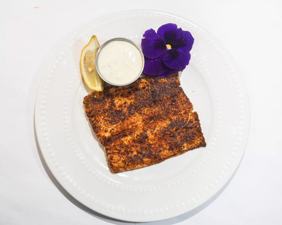 Blackened fish fillet with a lemon wedge, tartar sauce, and a purple edible flower on a white plate.