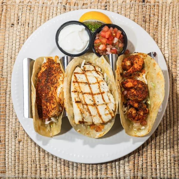Three tacos, one fish, one grilled fish, and one shrimp, with sides of sour cream and salsa.