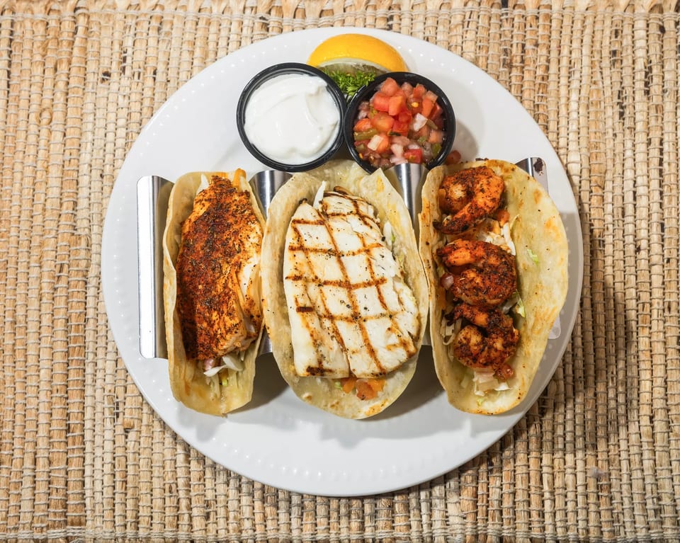 Three tacos, one fish, one grilled fish, and one shrimp, with sides of sour cream and salsa.