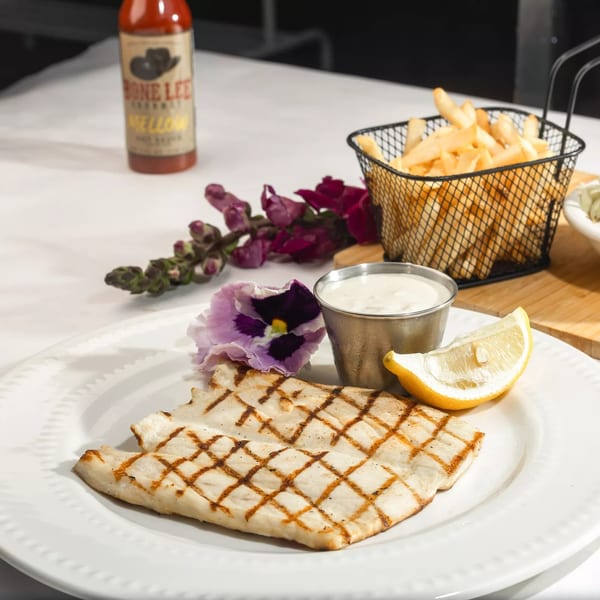 Grilled fish with fries, coleslaw, lemon, sauce, and a hot sauce bottle on a white table.