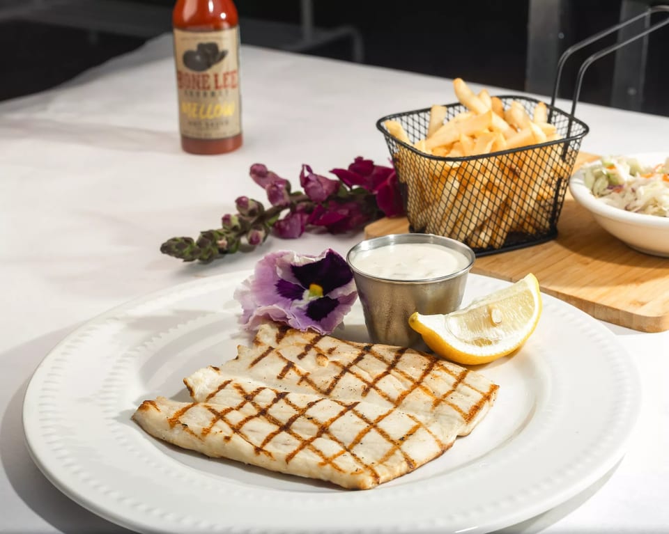 Grilled fish with fries, coleslaw, lemon, sauce, and a hot sauce bottle on a white table.